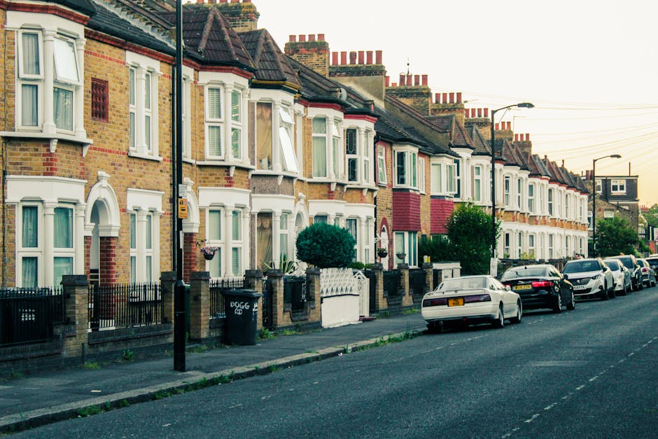 Photograph of a residential street in IG2 Ilford featuring a row of traditional terraced houses with bay windows, brick facades, and chimneys, illuminated by warm evening light. In front of the houses