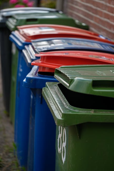 A row of large outdoor wheelie bins with closed lids, positioned along a sidewalk adjacent to a brick wall. The bins are in different colors, including green, blue, red, and dark green, and are made o