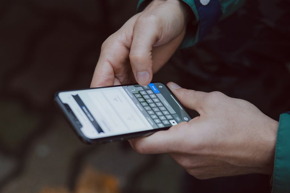 Close-up of a person’s hands holding a smartphone outdoors, with one hand supporting the device and the other tapping the screen, which displays an online form or quote interface. The individual is we